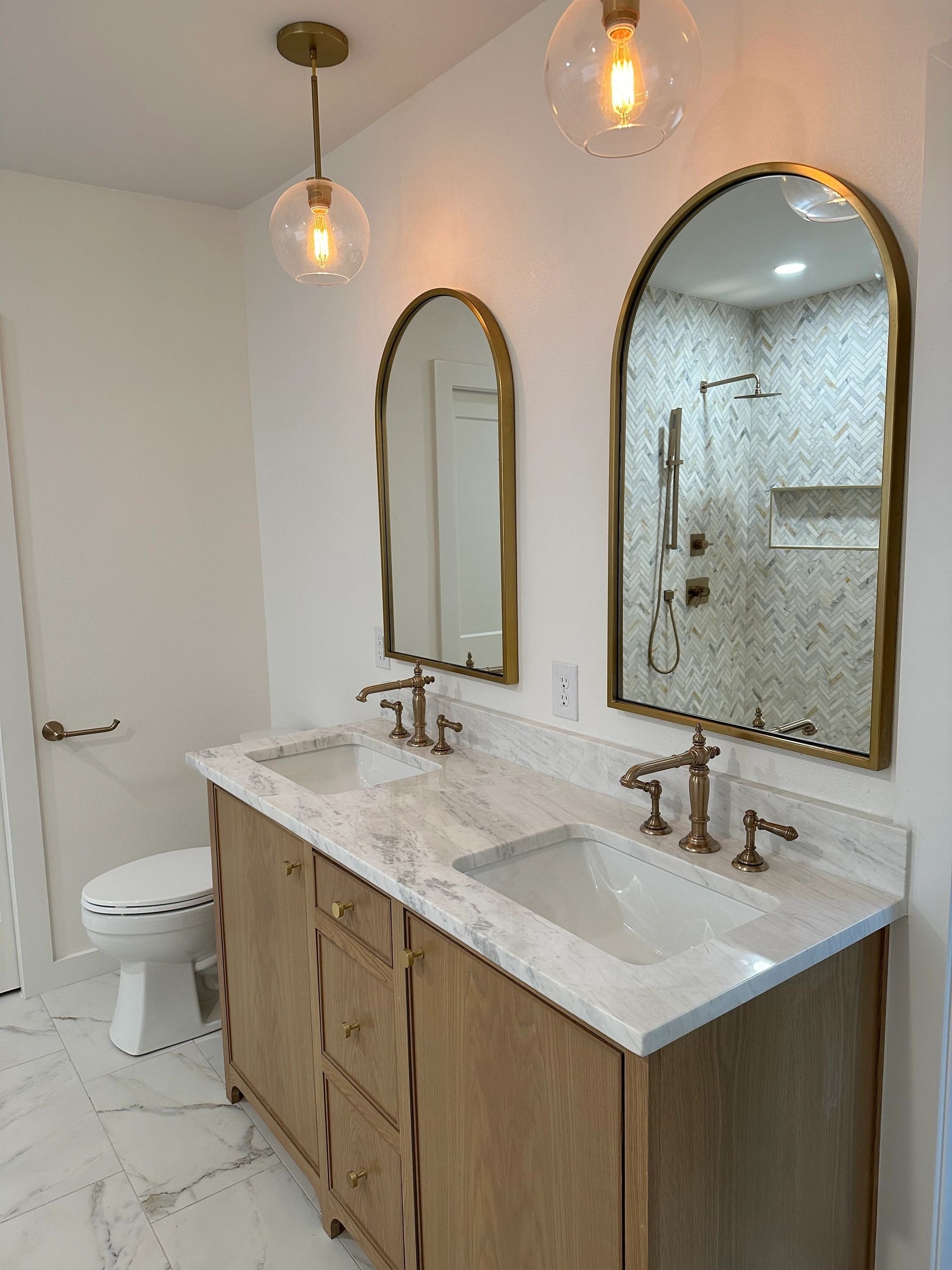 Custom bathroom with double wood vanity, brass arched mirrors, pendant lighting, and herringbone tile shower by BMW Renovations